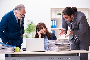 Woman on phone at desk being overloaded wiith work by two male colleagues